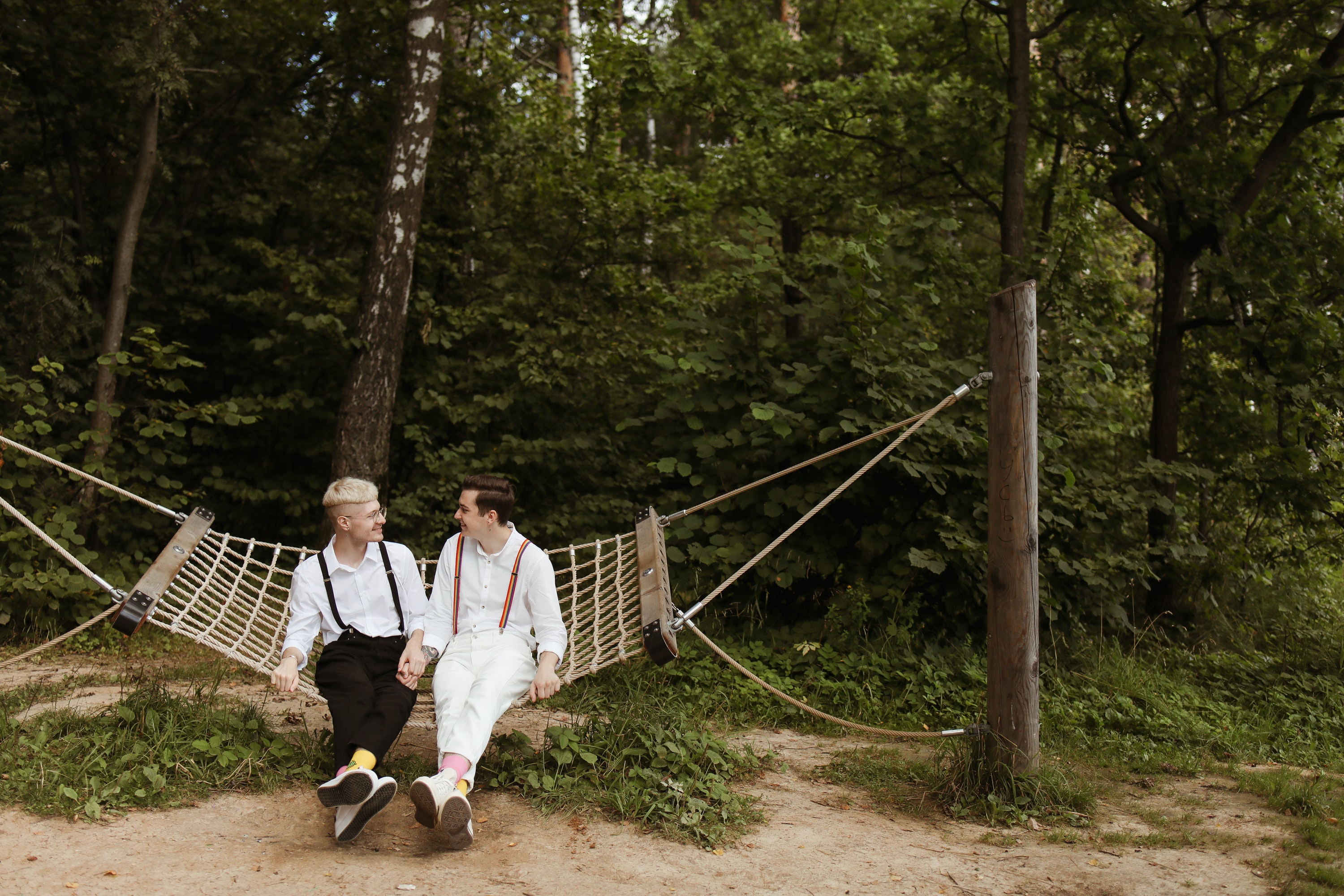grooms smiling at each other holding hands on a hammock with a green wooded backdrop
