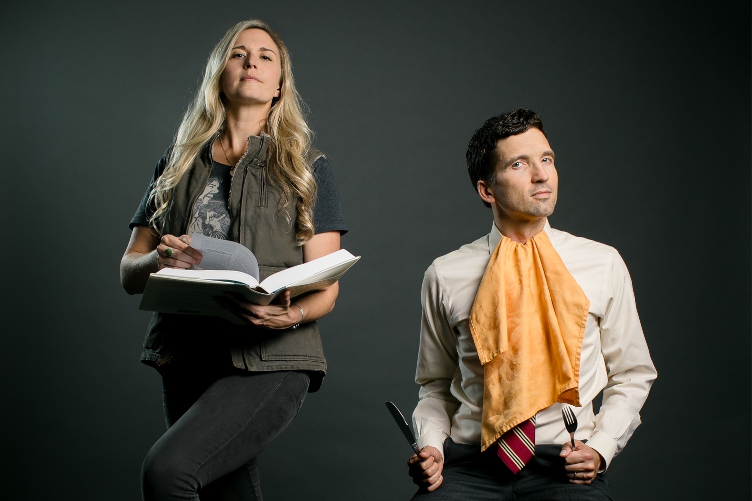 Lizzie Post (medium build with long blond hair) stands in casual attire - grey jeans, Bob Segar T-Shirt and Carhart Vest - holds open large etiquette book with her nose in the air. Daniel Post Senning (lean with brown hair) in a white button down shirt and yellow napkin tucked into his collar, is seated gripping a fork in his left fist and knife in his right. His head is cocked and his expression ever so slightly threatening. It is a humorous depiction of the two cousins who teach and write about etiquette